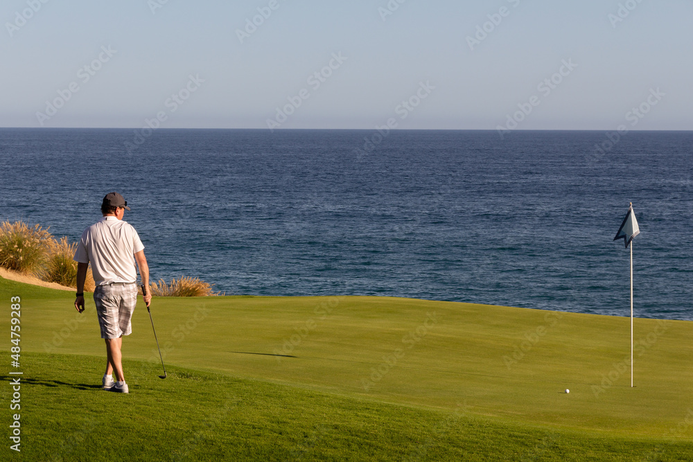 Middle aged man watching golf ball roll on golf green towards hole. Oceanside tropical golf course with blue ocean and clear blue sky in background. Copy space.