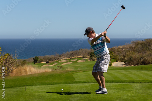 Middle aged man golfing, caught mid swing off the tee box. On ocean side tropical golf course with deep blue sea and clear blue sky as background. Copy space.