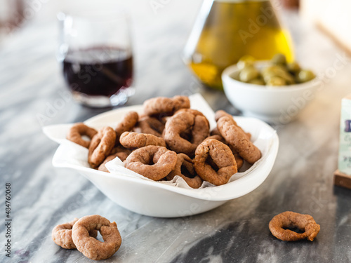 Taralli with red wine in a white bowl. Vegan aperitif. Glass of wine, olive oil and green olives in the background