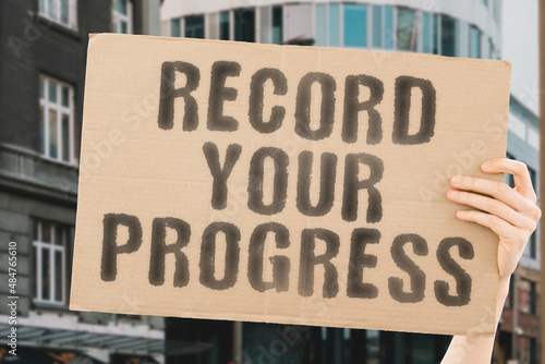 The phrase " Record your progress " on a banner in men's hand with blurred background. Rise. Increase. Improve. Evolution. Achieved. Level up. Updated. Updating. Tracking. Working. Worker. Time
