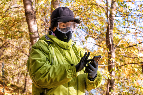 A woman wearing a respirator mask and protective glasses using a smartphone walking in the forest at COVID-19 period.