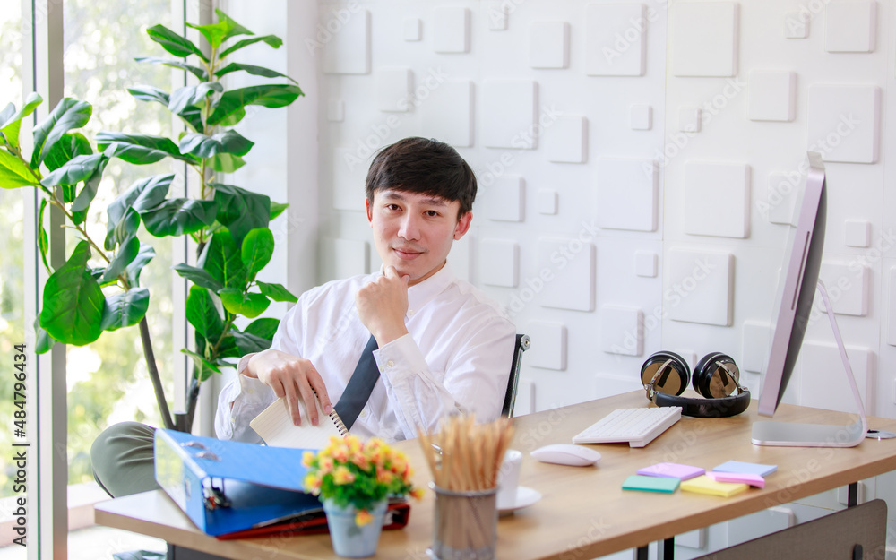 Portrait studio shot of Asian professional successful male businessman employee in formal shirt with necktie sitting look at camera at working desk with computer monitor keyboard mouse and stationery Portrait studio shot of Asian professional successful male businessman employee in formal shirt with necktie sitting look at camera at working desk with computer monitor keyboard mouse and stationery