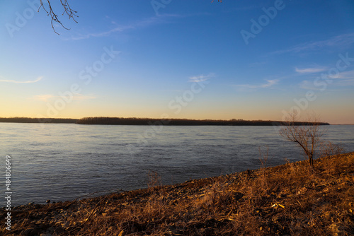 a stunning shot of the blue running waters of the Mississippi river with bare trees and brown and yellow grass along the banks of the river with blue sky and clouds at sunset at Greenbelt Park	 photo
