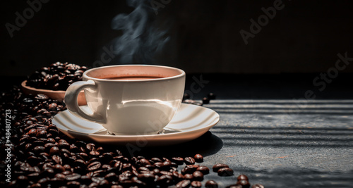 Hot coffee in a white coffee cup and many coffee beans placed around a on dark background.