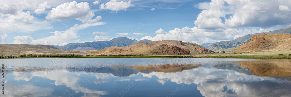 summer panorama of a mountain range with textured clouds and reflection in a mountain lake during the day in sunny weather