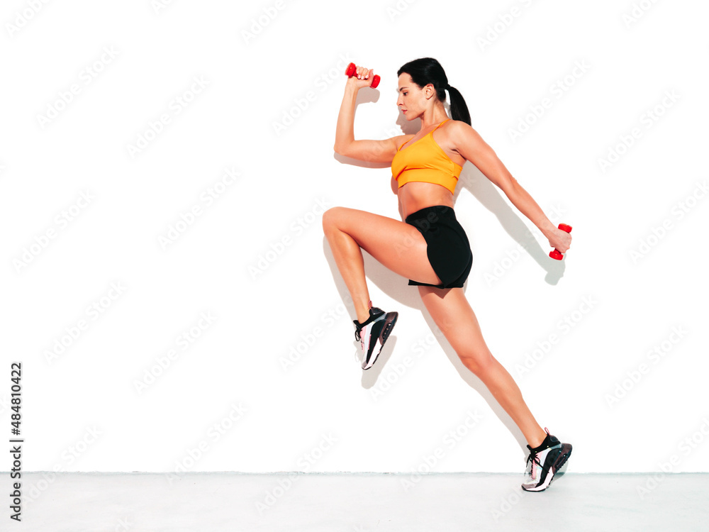 Fitness woman in sexy sports clothing. Young beautiful model with perfect body. Female posing in studio near white wall at summer sunny day. Jumping and running. Doing exercises with dumbbells