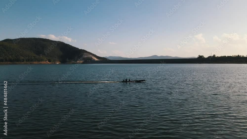 Long narrow traditional Thai boat traveling at great speed over Mae Ngat Somboonwith the Chon Dam in the background on a sunny day. Drone pedestal shot Long narrow traditional Thai boat traveling at great speed over Mae Ngat Somboonwith the Chon Dam in the background on a sunny day. Drone pedestal shot