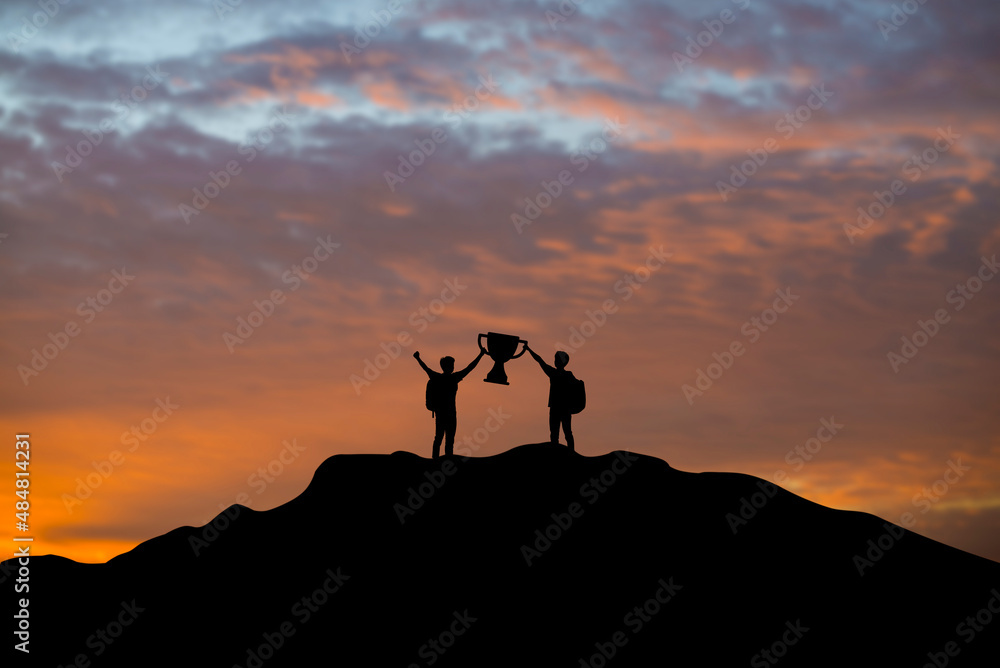 Silhouette of two young businessmen hold trophies on top of a mountain at sunset. Both of them demonstrated success in business and leadership.