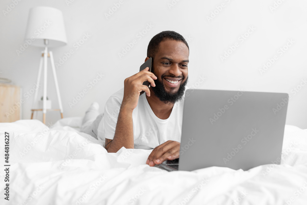 Smiling millennial black bearded guy lies on white bed in bedroom interior speaks by smartphone, looks at laptop Smiling millennial black bearded guy lies on white bed in bedroom interior speaks by smartphone, looks at laptop
