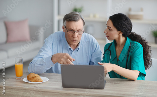 Young nurse showing senior man in wheelchair how to use laptop at retirement home photo