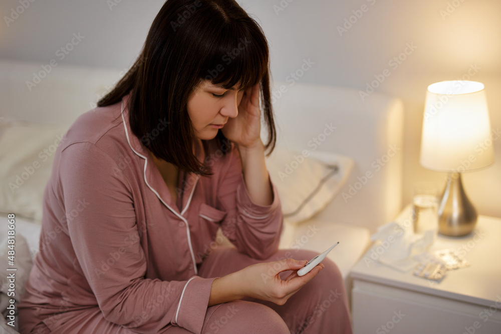 Sick young woman sitting on the bed with thermometer Sick young woman sitting on the bed with thermometer