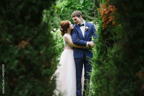 Bride and groom hugging behind the trees and foliage in the park on a green background, they smile and look at each other