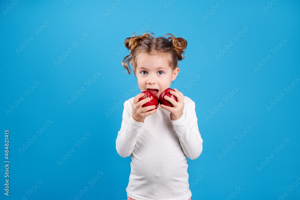 cute little girl with a funny hairstyle in a white T-shirt is holding two red apples in her hands. blue background. useful products for children. healthy snack. space for text. High quality photo cute little girl with a funny hairstyle in a white T-shirt is holding two red apples in her hands. blue background. useful products for children. healthy snack. space for text. High quality photo