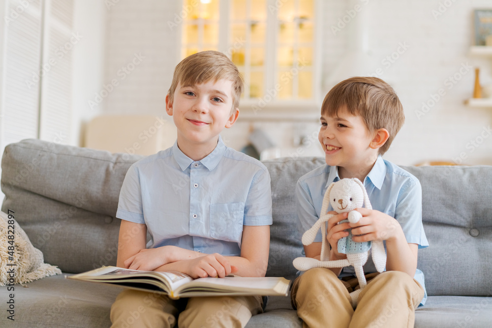Two boys sitting on couch are reading book. Older brother reads interesting stories to his younger brother, concept friendship, intelligence, interaction, younger brother holds toy rabbit in his hand Two boys sitting on couch are reading book. Older brother reads interesting stories to his younger brother, concept friendship, intelligence, interaction, younger brother holds toy rabbit in his hand