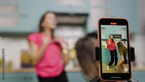 Two young girls dancing, sing, having fun and filming videos for their blogs or social networks. Bloggers are recording vlog with trend content in kitchen. Slow motion ready, 4K at 59.97fps. photo