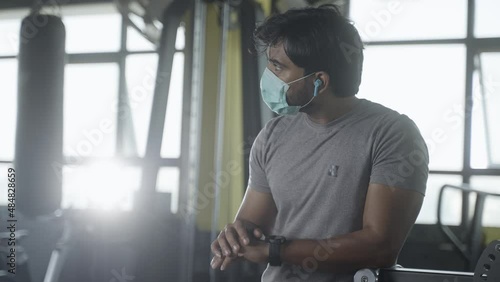 Young muscular builder with medical face mask checking workout plan or monitoring hart rate from smartwatch at gym - concpet of healthcare, technology and safety measures during covid-19 pandemic. photo