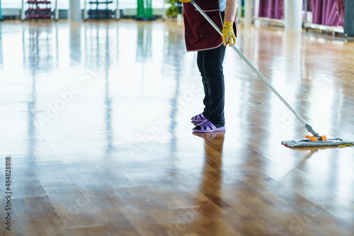 Unrecognizable cleaning woman in red uniform, yellow gloves, purple rubber slippers mopping linoleum floor in large empty room with sport equipment. Cleaning, disinfection, cleaning service concept. 