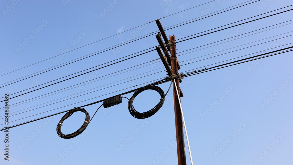 Black boxes and cable reels on poles. Internet box or splitter box with high-speed fiber optic cable hanging on a metal electric pole on a blue background with copy space. Selective focus - obrazy, fototapety, plakaty 