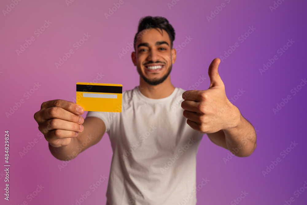 Happy young Arab man holding credit card and showing thumb up gesture in neon light, selective focus Happy young Arab man holding credit card and showing thumb up gesture in neon light, selective focus
