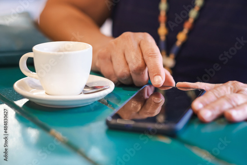 Addicted adult mature caucasian woman sitting at the table with white cup of coffee while texting with mobile phone. Unrecognizable female people enjoying social and technology using smartphone