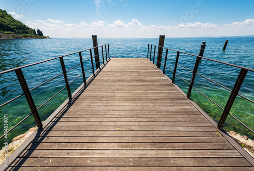 Small empty wooden pier on te coast of Lake Garda (Lago di Garda) in front of the small town of Garda, tourist resort in Verona province, Veneto, Italy, southern Europe.