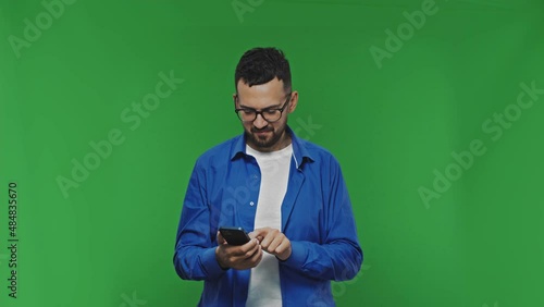 Smiling handsome bearded mand in casual shirt isolated on green background studio. Using mobile phone typing sms message browsing
