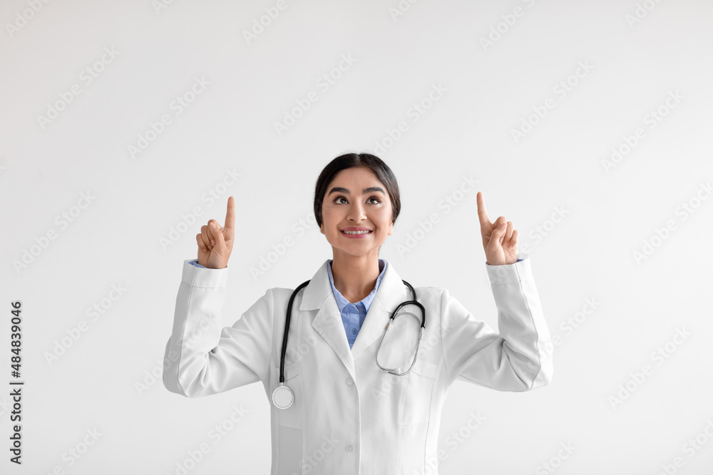 Smiling millennial hindu lady doctor in white coat with stethoscope shows fingers up to empty space Smiling millennial hindu lady doctor in white coat with stethoscope shows fingers up to empty space