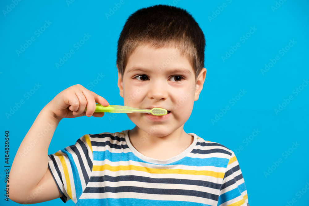Beautiful happy child in striped T-shirt brushes teeth with a green toothbrush on a blue background, copy space for text. Beautiful happy child in striped T-shirt brushes teeth with a green toothbrush on a blue background, copy space for text.
