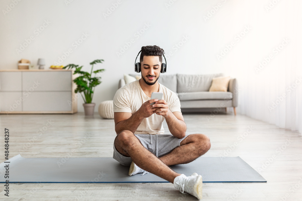 Fototapeta premium Young Arab guy in headset choosing music playlist for his domestic workout on mobile phone at home
