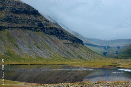A slope of an Icelandic glacier mountain with a lake in the foreground and clouds in the sky