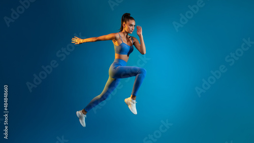 Fitness Woman Jumping In Mid-Air Over Blue Background, Panorama, Full-Length