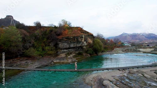 A girl on a suspension bridge to cross a mountain river with rocky shores and an autumn forest