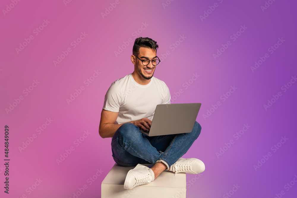 Full length of young Arab man sitting cross legged with laptop, having video call, working online in neon light Full length of young Arab man sitting cross legged with laptop, having video call, working online in neon light
