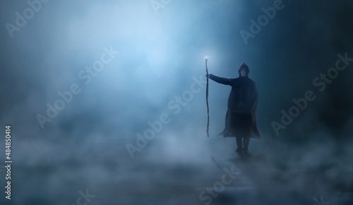 Magician in cloak, cowl with magic stick standing in fog landscape illuminated by blue moon ligh. Fantasy, wizard concept, 3D rendering