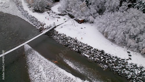 Top view of the mountain river and suspension bridge. People are standing on the snowy shore and trees are growing