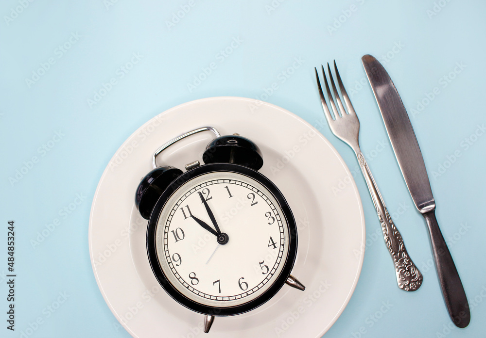 Concept of intermittent fasting, ketogenic diet, weight loss. fork and knife, alarmclock on plate Concept of intermittent fasting, ketogenic diet, weight loss. fork and knife, alarmclock on plate