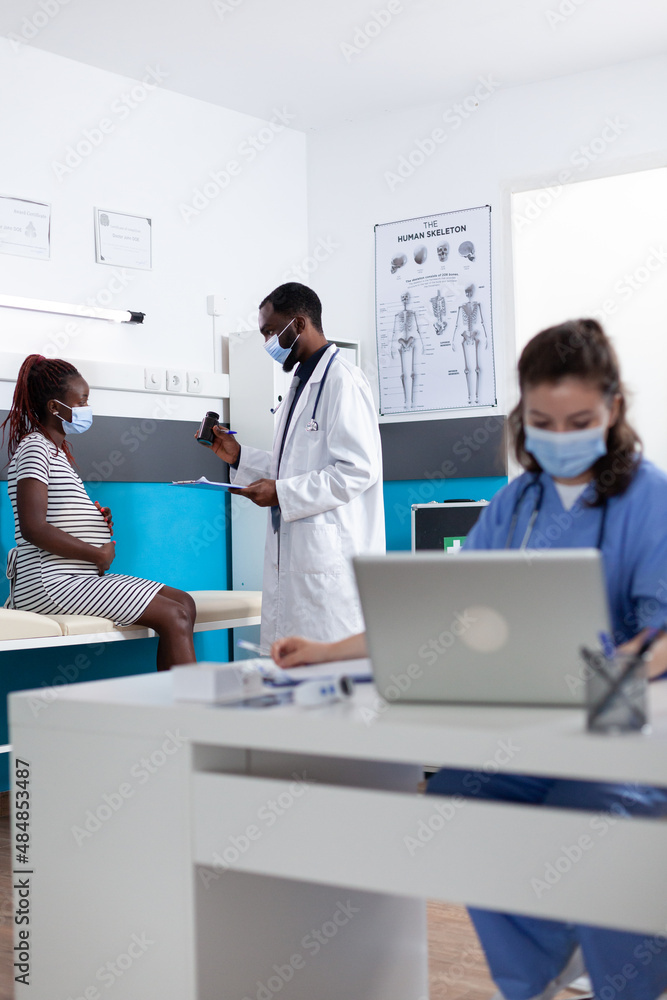 Doctor giving bottle of pills to pregnant woman at consultation. Pregnant patient talking to specialist at checkup visit, preparing to give birth to child during coronavirus pandemic.