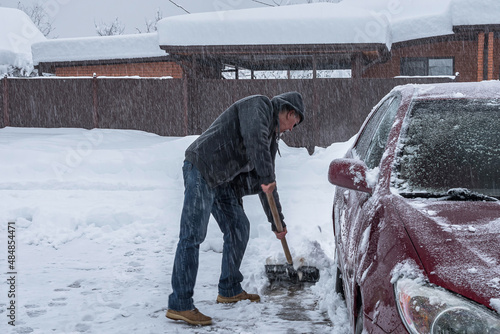 A man is clearing the street of snow with a shovel. A red car is standing nearby. In the background is a gray sky, snowy roofs of houses and snowfall. Clearing the street during a snow storm.