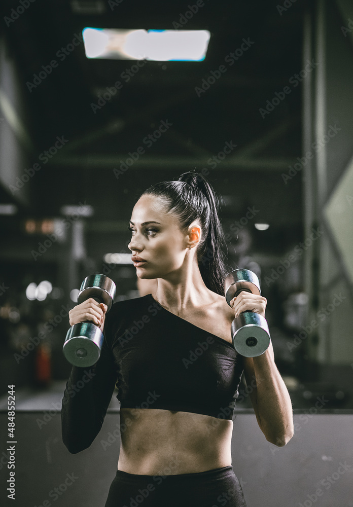 Fashionable young woman in a sports top with dumbbells in her hands on the background of the gym