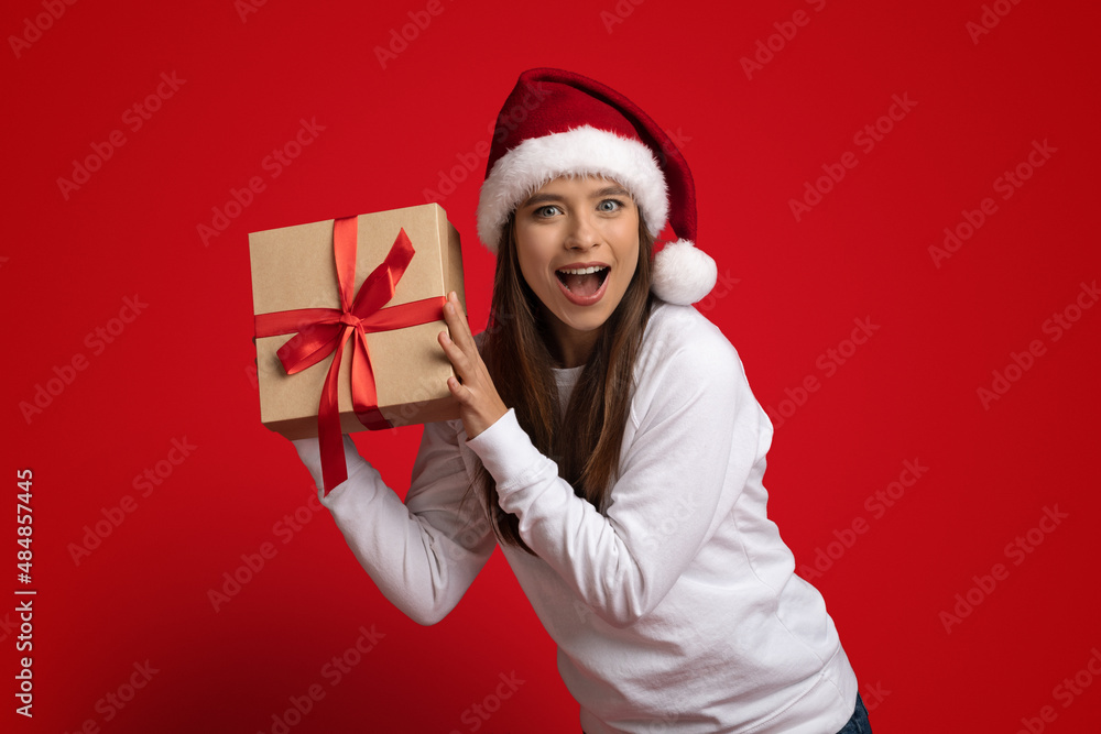 Holiday Bonus. Excited Young Woman Wearing Santa Hat Holding Wrapped Gift Box Holiday Bonus. Excited Young Woman Wearing Santa Hat Holding Wrapped Gift Box