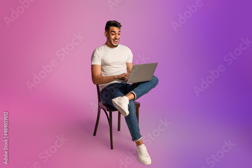 Shocked young Arab guy looking at laptop screen, sitting on chair, excited about online win or success in neon light