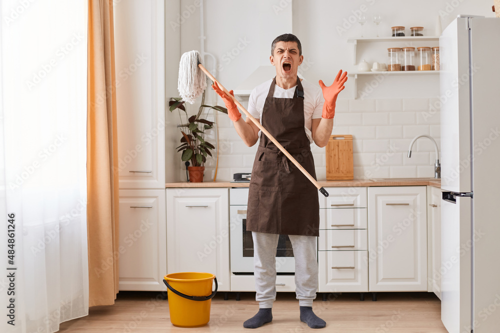 Indoor shot of male housekeeper wearing white t shirt and brown apron, standing in kitchen with mop in hands and screaming, being extremely tired washing his apartment. Indoor shot of male housekeeper wearing white t shirt and brown apron, standing in kitchen with mop in hands and screaming, being extremely tired washing his apartment.
