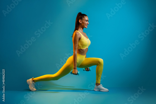 Sporty Female Doing Deep Lunge Exercise Holding Dumbbells, Blue Background