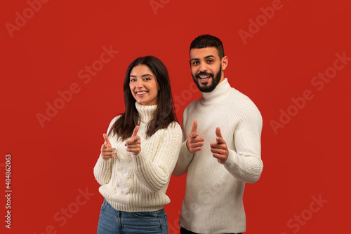 Your turn. Happy middle eastern couple pointing at camera with fingers, picking somebody, posing over red background photo