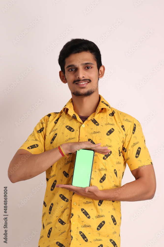 Image of unshave attractive indian guy with wearing yellow shirt and showing cell phone on camera and gesturing cool sign with hand isolated on white background studio portrait Image of unshave attractive indian guy with wearing yellow shirt and showing cell phone on camera and gesturing cool sign with hand isolated on white background studio portrait