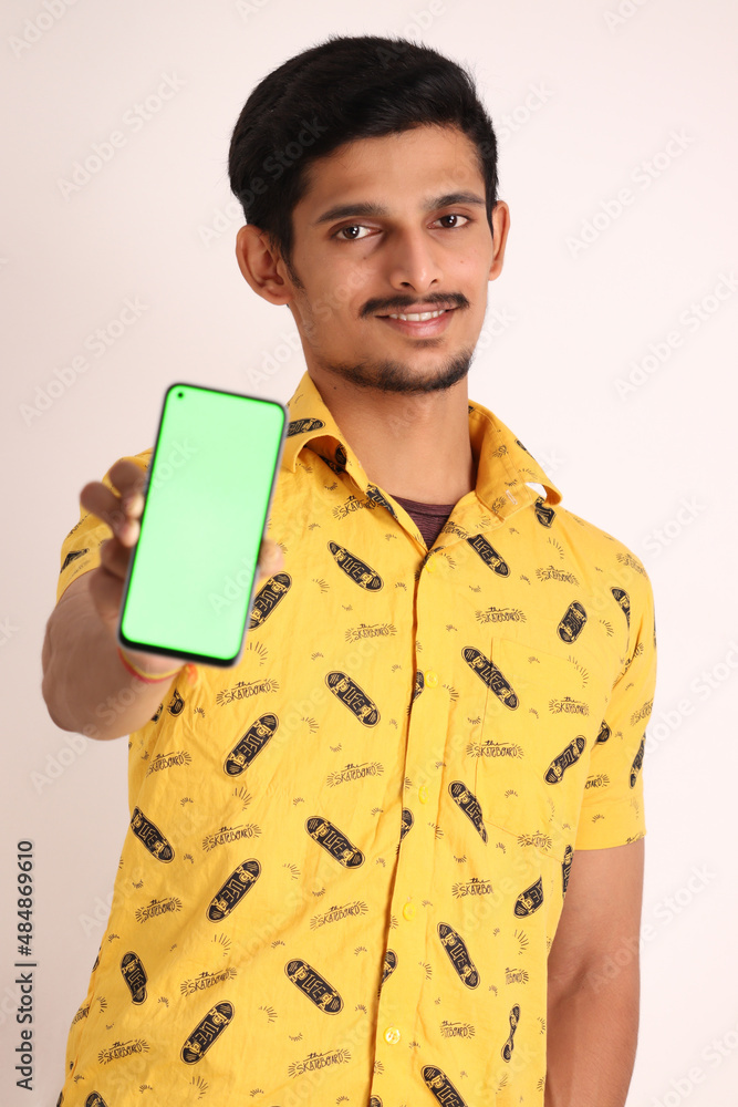 Image of unshave attractive indian guy with wearing yellow shirt and showing cell phone on camera and gesturing cool sign with hand isolated on white background studio portrait Image of unshave attractive indian guy with wearing yellow shirt and showing cell phone on camera and gesturing cool sign with hand isolated on white background studio portrait