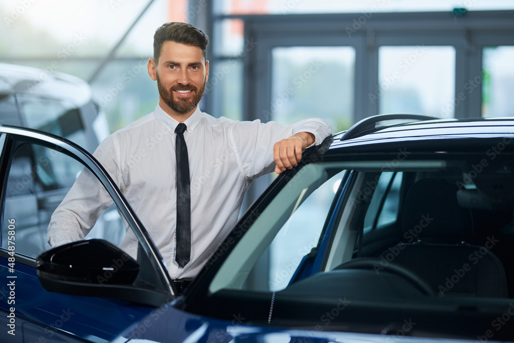 Handsome bearded man in formal outfit visiting auto salon for choosing new vehicle. Happy male customer standing near open door of luxury car and smiling on camera. 