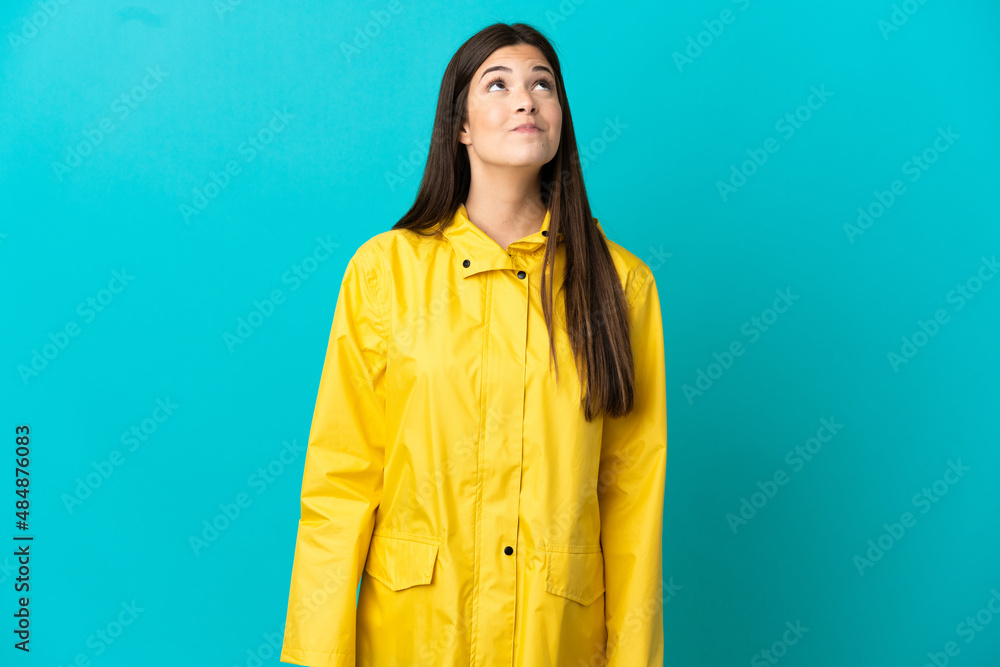 Teenager Brazilian girl wearing a rainproof coat over isolated blue background and looking up Teenager Brazilian girl wearing a rainproof coat over isolated blue background and looking up