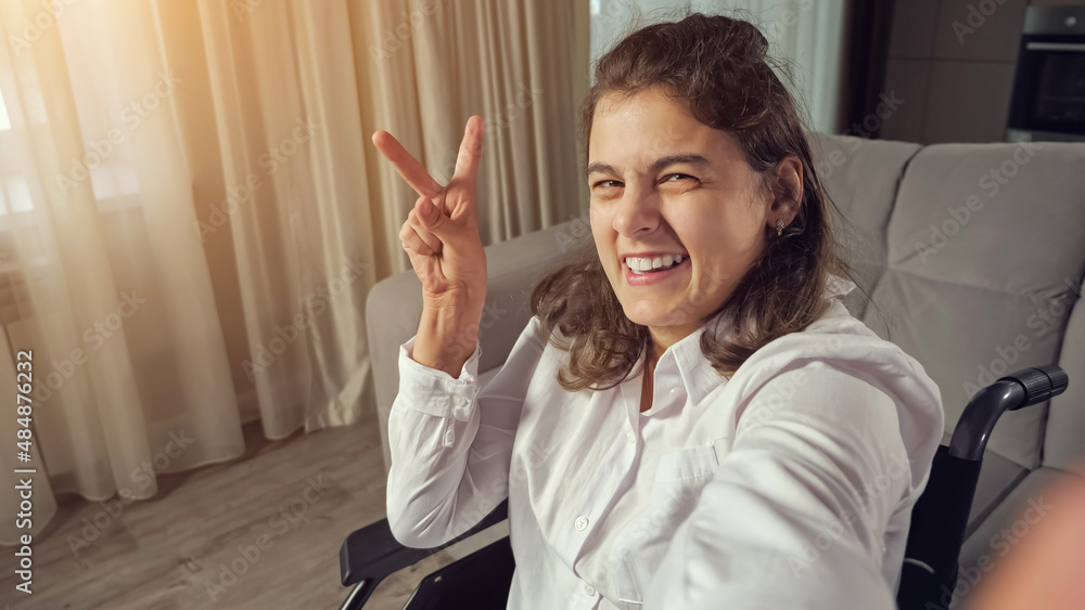 Disabled woman with cerebral palsy makes fingers Victory sign and smiles waving hand sitting in wheelchair expressing equality of people with disabilities. - obrazy, fototapety, plakaty 