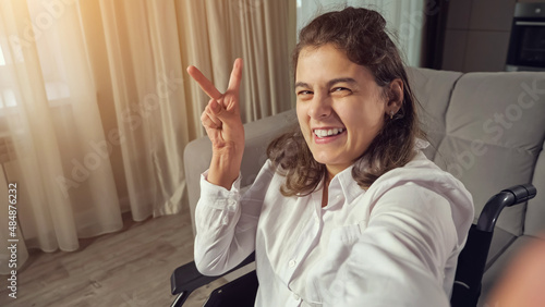 Disabled woman with cerebral palsy makes fingers Victory sign and smiles waving hand sitting in wheelchair expressing equality of people with disabilities. photo
